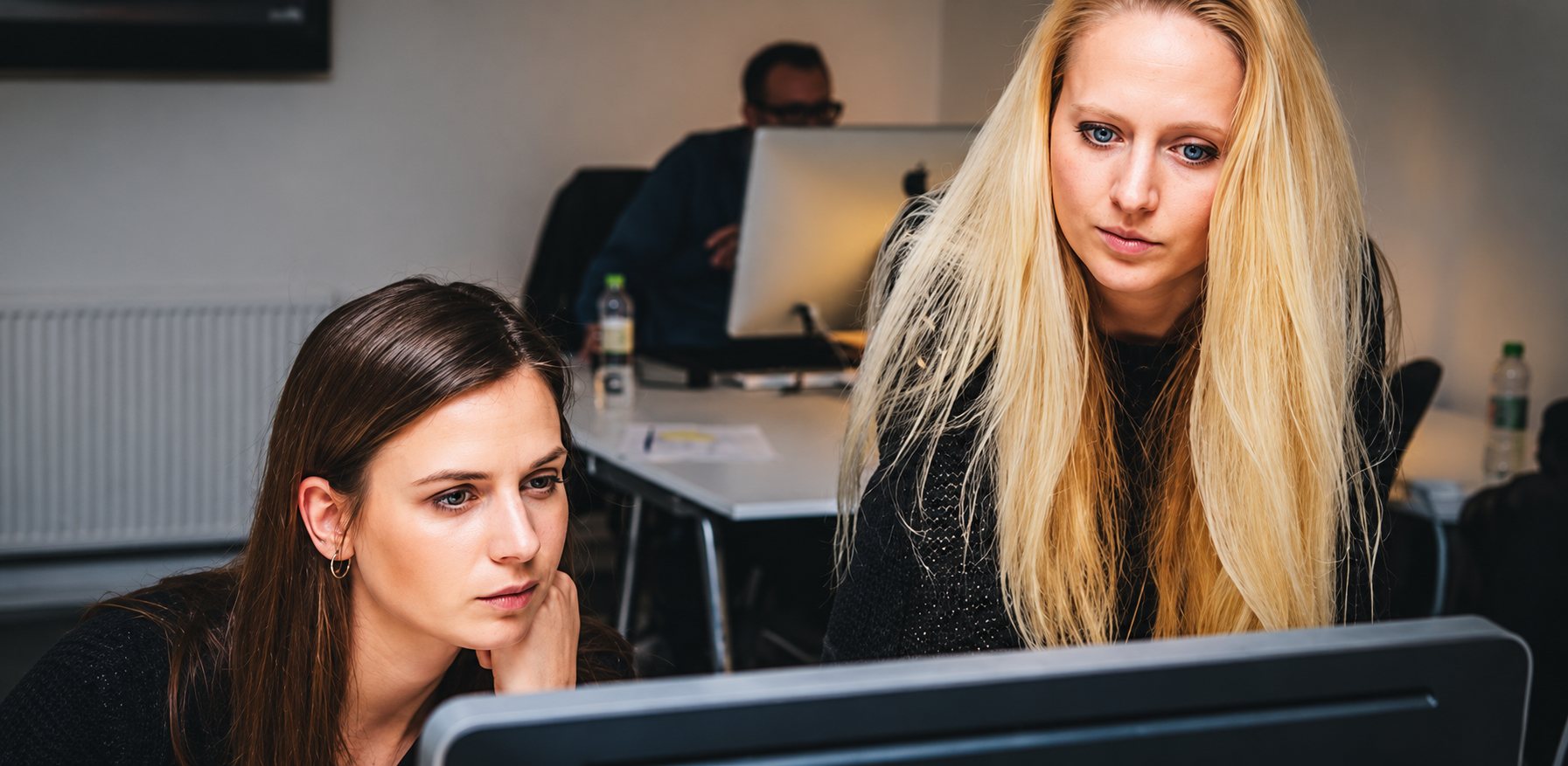 Deux jeunes femmes, l'une brune et l'autre blonde, regardent attentivement un écran d'ordinateur dans un bureau où un collègue travaille en arrière-plan.