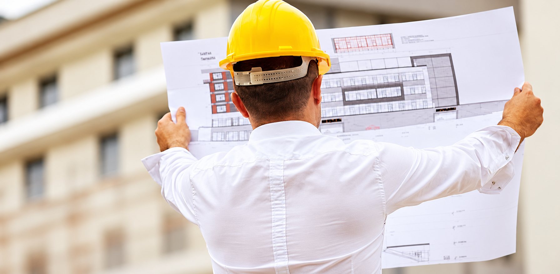 Un homme de dos, vêtu d'une chemise blanche et coiffé d'un casque de chantier jaune, déplie un grand plan de bâtiment devant une façade floue.
