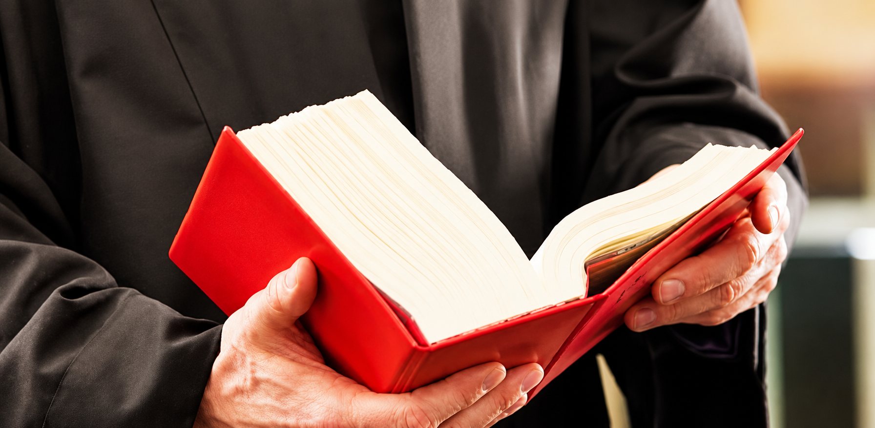 Une personne vêtue d'une robe sombre tient entre ses mains un gros livre à couverture rouge ouvert.